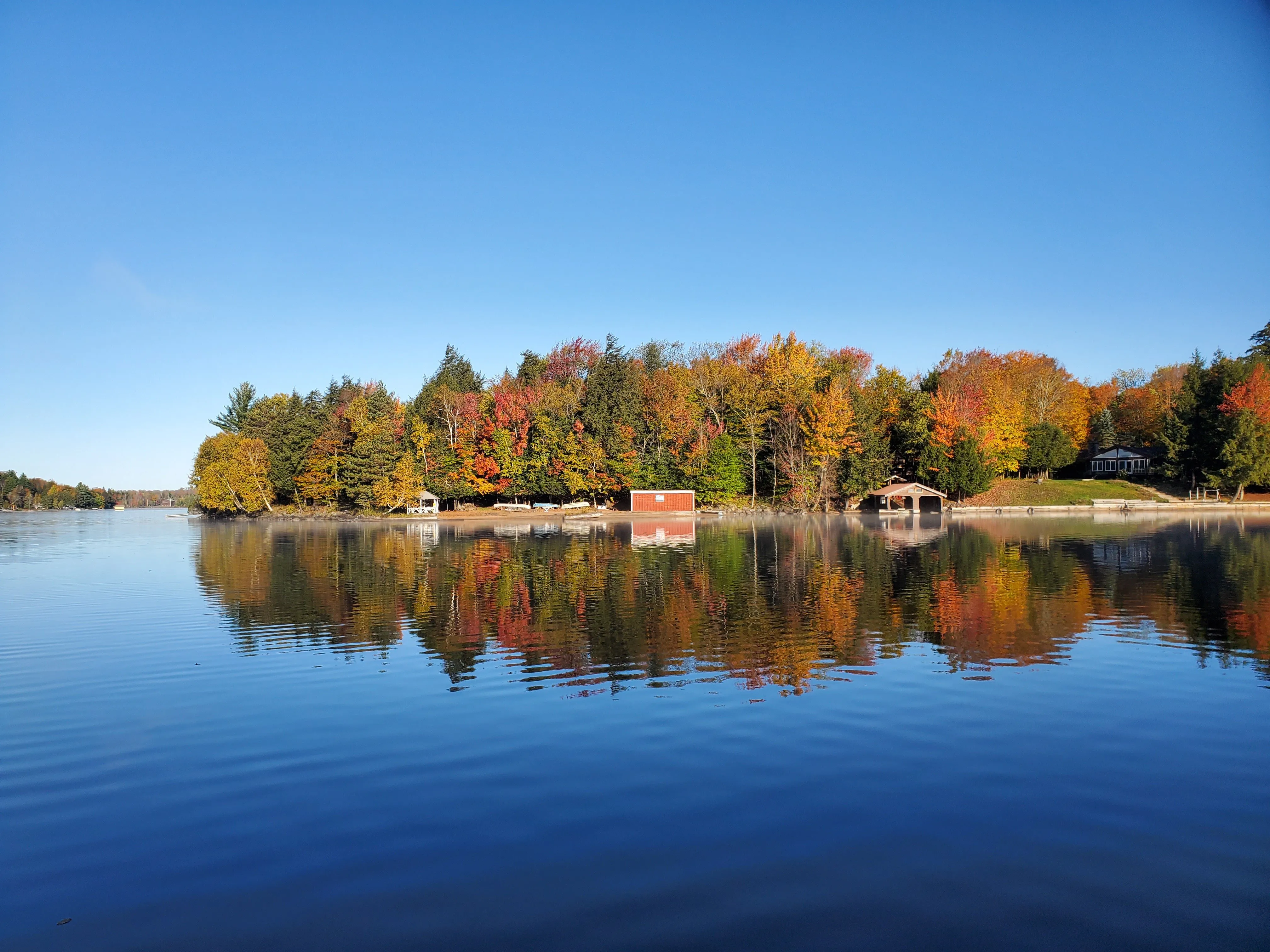 A calm autumn morning on Cranberry Lake, with a fall-colored shoreline and a small red cabin reflected in glass-still water under a clear blue sky.