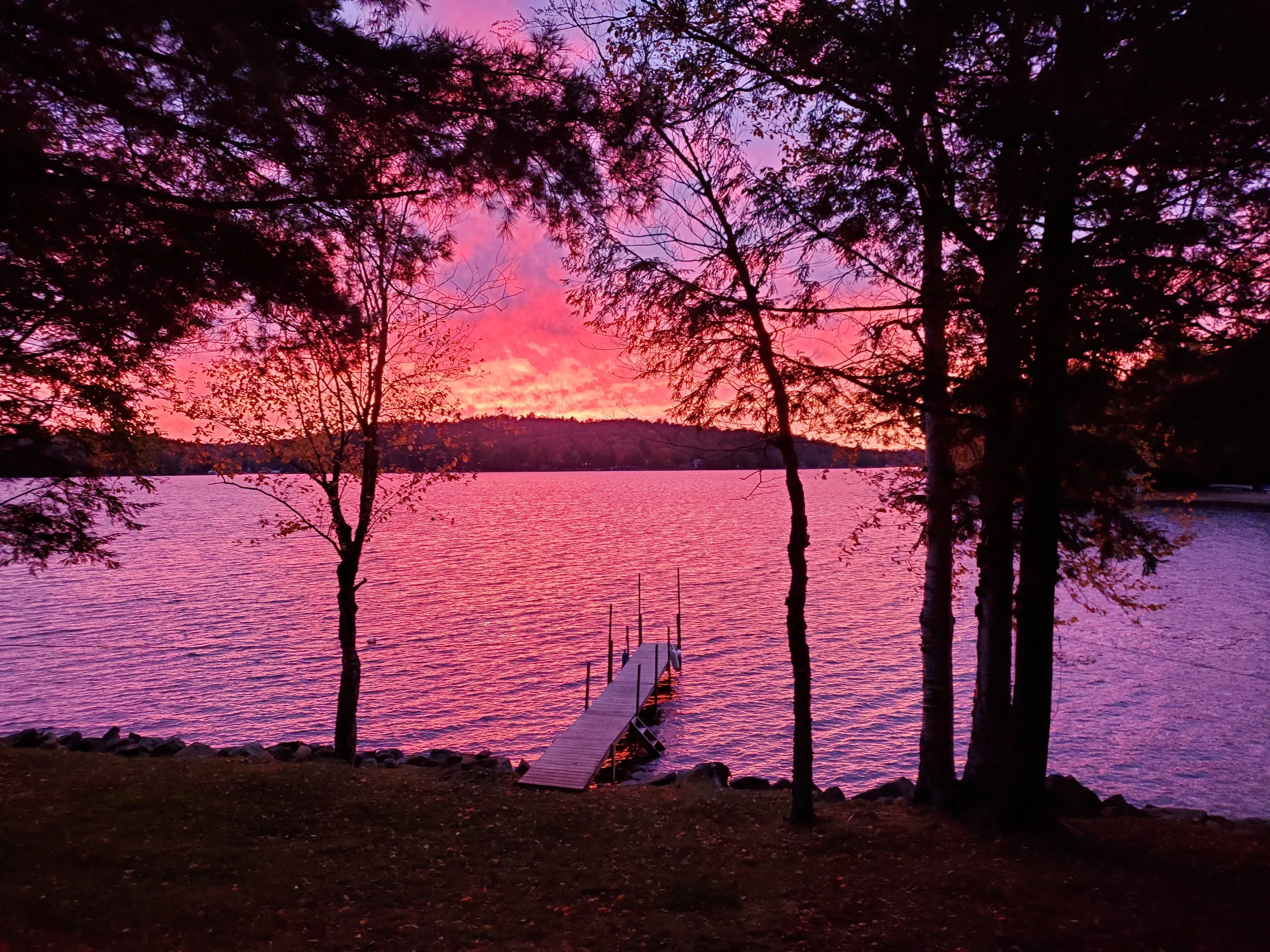 A vivid pink and magenta sunset over Cranberry Lake, framed by silhouetted trees, with the dock reaching out into glowing water.