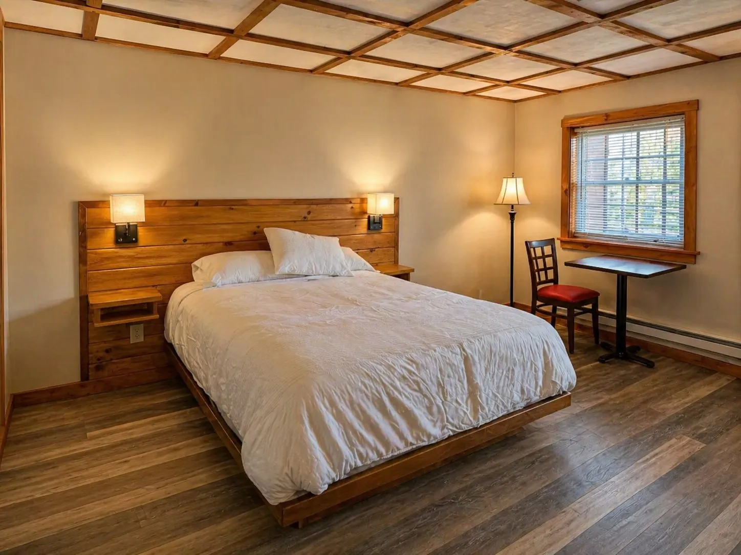 Newly renovated motel room — custom wood-slat headboard, white bedding, lamp-lit reading nook, and a small writing desk by the window.